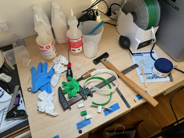 a messy workbench with a 3d printer spool, resin bottles, gloves, a hammer, a clamp, demolding detritus, and a powertowel with some blood on it (not too much, just like a little cut that you didn't have bandaids for but also wouldn't stop bleeding).